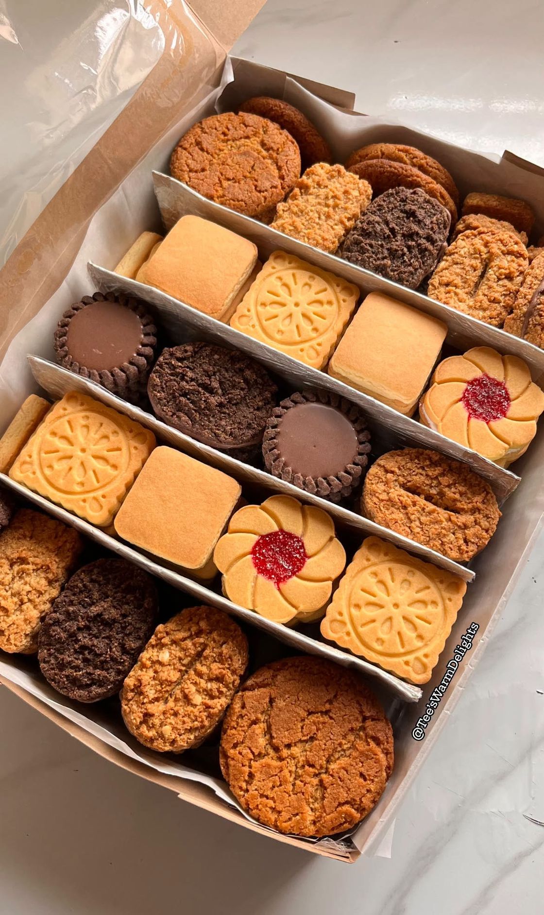 An open box of assorted homemade biscuits — shortbread, chocolate creams, ginger snaps and jam whirls — packed in neat rows.